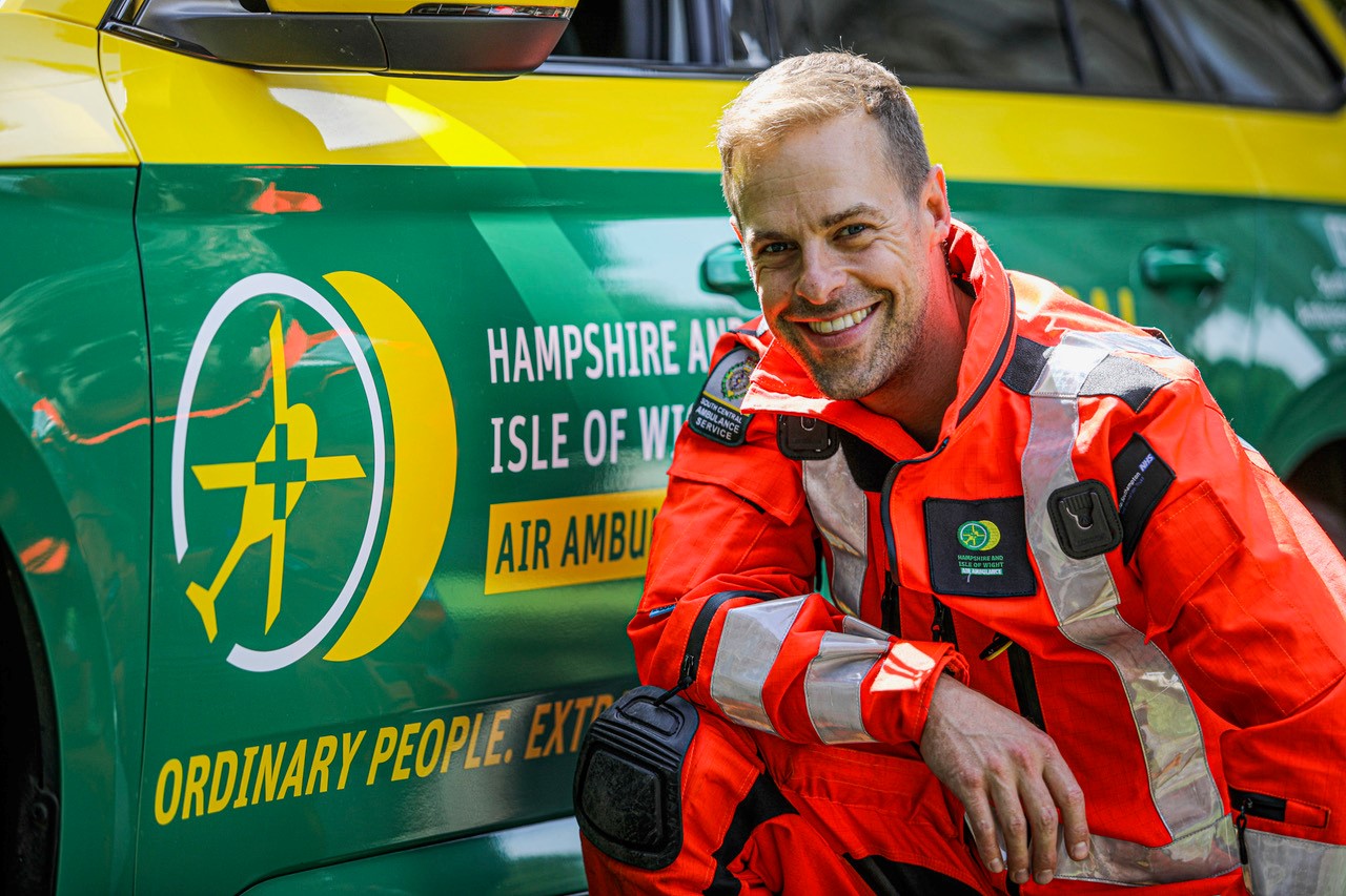 Dr Jamie Plumb is crouched in front of the HIOWAA emergency response vehicle and smiling to the camera. He is wearing the full flight suit.